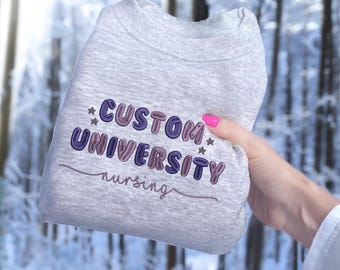May include: A gray sweatshirt with the text "CUSTOM UNIVERSITY nursing" embroidered in purple and pink letters with stars on either side.