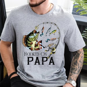 May include: A gray t-shirt with a graphic of a fish on a fishing hook. The text "Hooked on Papa" is printed below the fish. The names "Jace", "Kaleb", "Jamison", "Brantley", and "Paxton" are printed on the fishing lures.