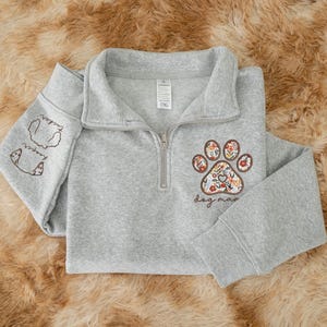 May include: A gray quarter-zip sweatshirt featuring a floral paw print design and the words "dog mom." The left sleeve has dog illustrations and the names "Tucker" and "Harper." The sweatshirt has a ribbed collar and cuffs.