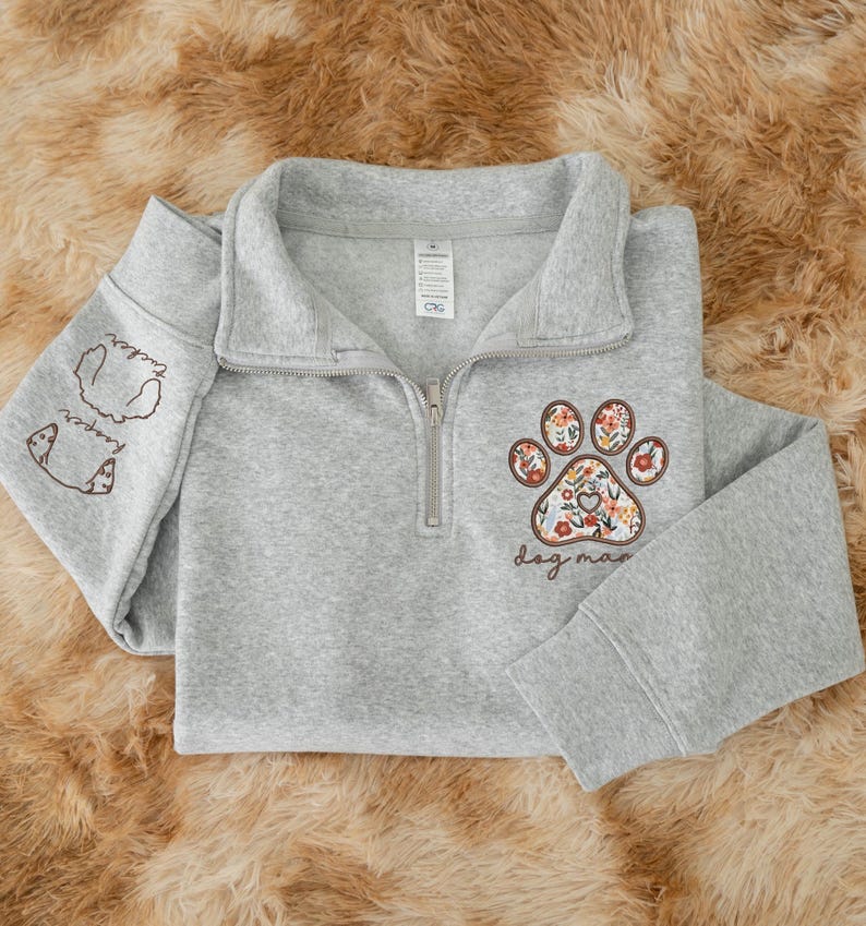 May include: A gray quarter-zip sweatshirt featuring a floral paw print design and the words "dog mom." The left sleeve has dog illustrations and the names "Tucker" and "Harper." The sweatshirt has a ribbed collar and cuffs.