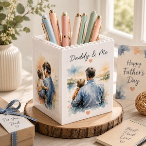 May include: A white square pen holder with a building block texture on the top rim. The sides feature watercolor illustrations of a parent and child with the text Daddy & Me. Several pastel-colored pens are stored inside the container.