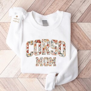 Op de afbeelding: Witte sweatshirt met de tekst "CORSO MOM" in een bloemenontwerp. De letters zijn gevuld met een patroon van rode, oranje en gele bloemen en groene bladeren. De mouw heeft de tekst "Dublin Cooper".