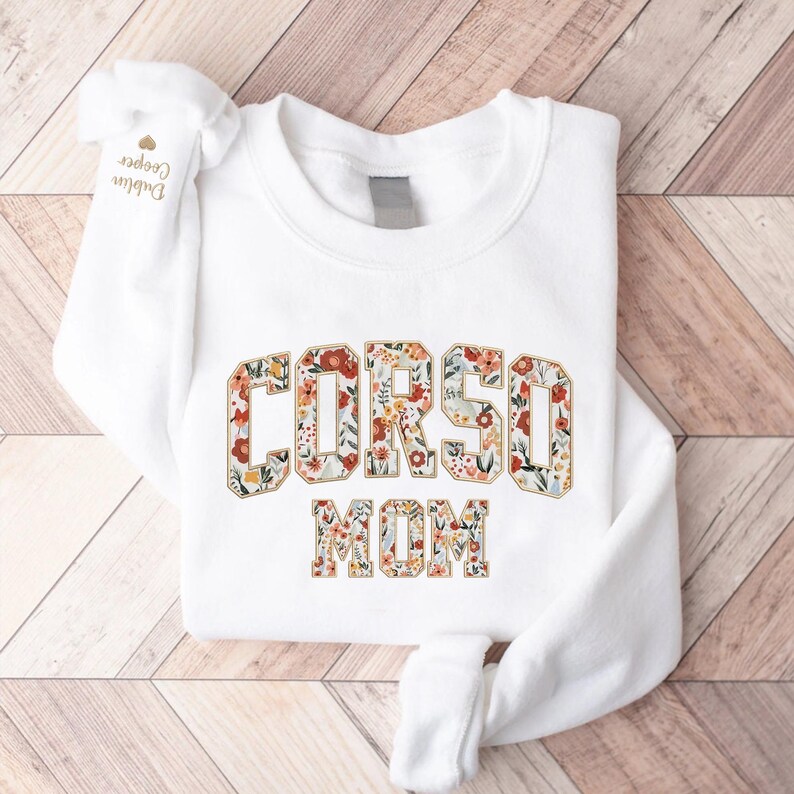 Op de afbeelding: Witte sweatshirt met de tekst "CORSO MOM" in een bloemenontwerp. De letters zijn gevuld met een patroon van rode, oranje en gele bloemen en groene bladeren. De mouw heeft de tekst "Dublin Cooper".