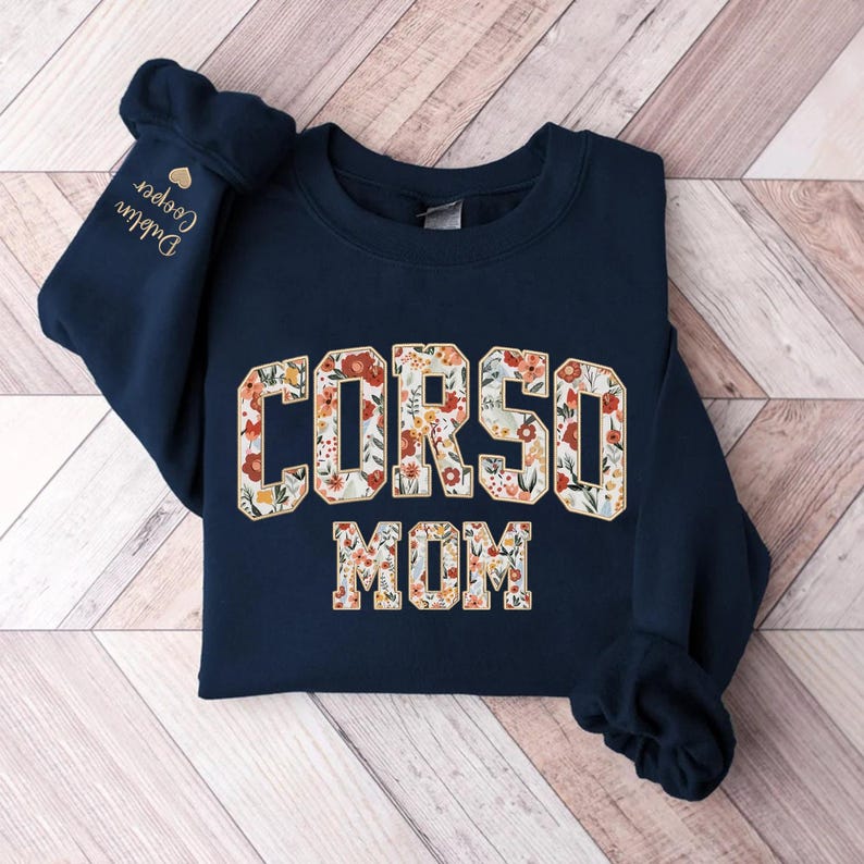 Op de afbeelding: Marineblauw sweatshirt met de tekst "CORSO MOM" in een bloemenpatroon. De linkermouw toont de naam "Dublin Cooper" in gouden borduursel, samen met een gouden hart.