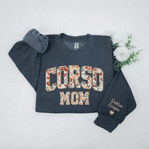 Op de afbeelding: Een donkergrijs sweatshirt met de tekst "CORSO MOM" in een bloemenpatroon. De mouw toont de naam "Dublin Cooper" en een hart. Het sweatshirt ligt op een witte ondergrond.