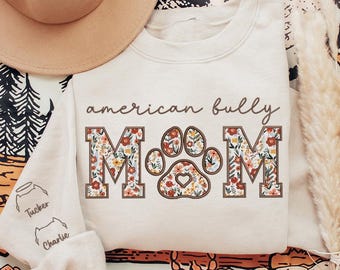 May include: Cream-colored sweatshirt with the text "american bully mom" in a brown script. The word "MOM" is in a floral design with a paw print in the center. The sleeve has the names "Tucker" and "Charlie" with dog illustrations.