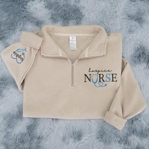 May include: A beige quarter-zip sweatshirt and matching hat. The sweatshirt has the words "hospice NURSE" printed in black and blue, with a stethoscope graphic. The hat is embroidered with the name "Sophia" and a stethoscope.