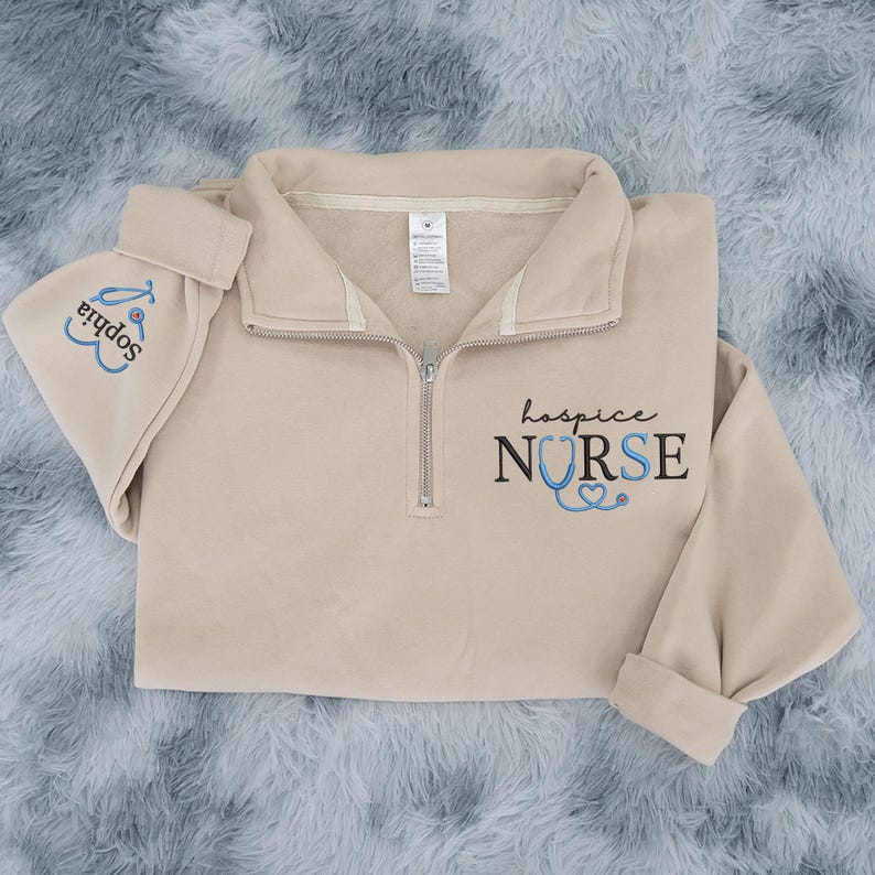 May include: A beige quarter-zip sweatshirt and matching hat. The sweatshirt has the words "hospice NURSE" printed in black and blue, with a stethoscope graphic. The hat is embroidered with the name "Sophia" and a stethoscope.