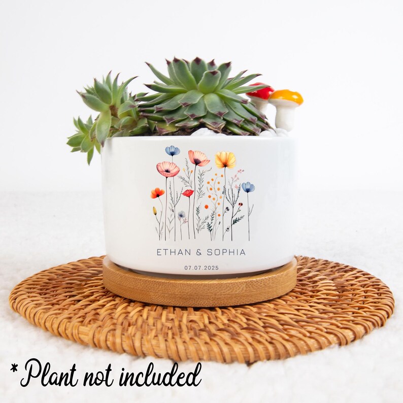 May include: White ceramic planter with a floral design and the text "ETHAN & SOPHIA 07.07.2025". The planter sits on a round, woven, brown placemat. A succulent plant and decorative mushrooms are inside the planter.