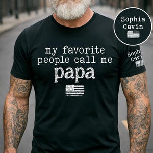 May include: Black t-shirt with the text "my favorite people call me papa" in white, along with an American flag graphic. The shirt also features the name "Sophia Cavin" and a small American flag on the sleeve.