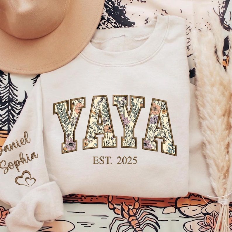 May include: Cream-colored sweatshirt with the word "YAYA" in a floral design, outlined in brown. Below the word is "EST. 2025". The sleeve on the left has the names "Daniel Sophia" and a heart symbol. A tan hat is visible in the upper left.