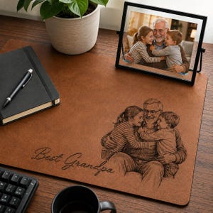May include: A brown desk mat with a laser-etched image of a grandfather hugging two grandchildren. The words "Best Grandpa" are etched on the mat. A framed photo of the same image is on the desk, along with a black notebook and pen.