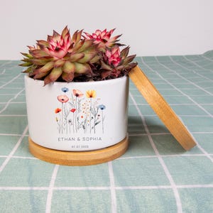 May include: A white ceramic planter with a floral design and the names "ETHAN & SOPHIA" and the date "07.07.2025". The planter holds a succulent plant with red and green leaves. It sits on a round bamboo base with a matching bamboo tray.