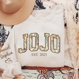 May include: Cream-colored sweatshirt with the word "JOJO" in a floral design. The text "EST. 2025" is below the word. A tan hat is in the upper left corner. The sleeve on the left has the names "Daniel Sophia" and a heart.