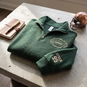 May include: A dark green quarter-zip sweatshirt featuring gold embroidery on the chest with a football graphic and the text football mom. The sleeve is embroidered with the name ETHAN and the number 99 in gold thread, matching the chest design.