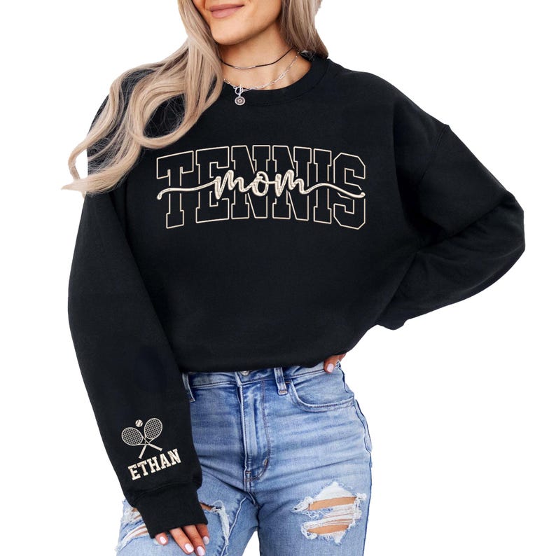 May include: Black sweatshirt with the text "TENNIS mom" in a white outlined font. The sleeve features a tennis racket and ball graphic with the name "ETHAN" below it. The sweatshirt is worn with blue jeans.