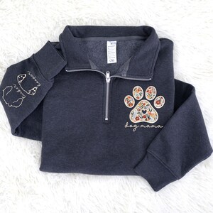 May include: Dark gray quarter-zip sweatshirt with a floral paw print design and the words "dog mama." The left sleeve features embroidered dog silhouettes and names. The sweatshirt has a silver zipper and a soft, comfortable appearance.