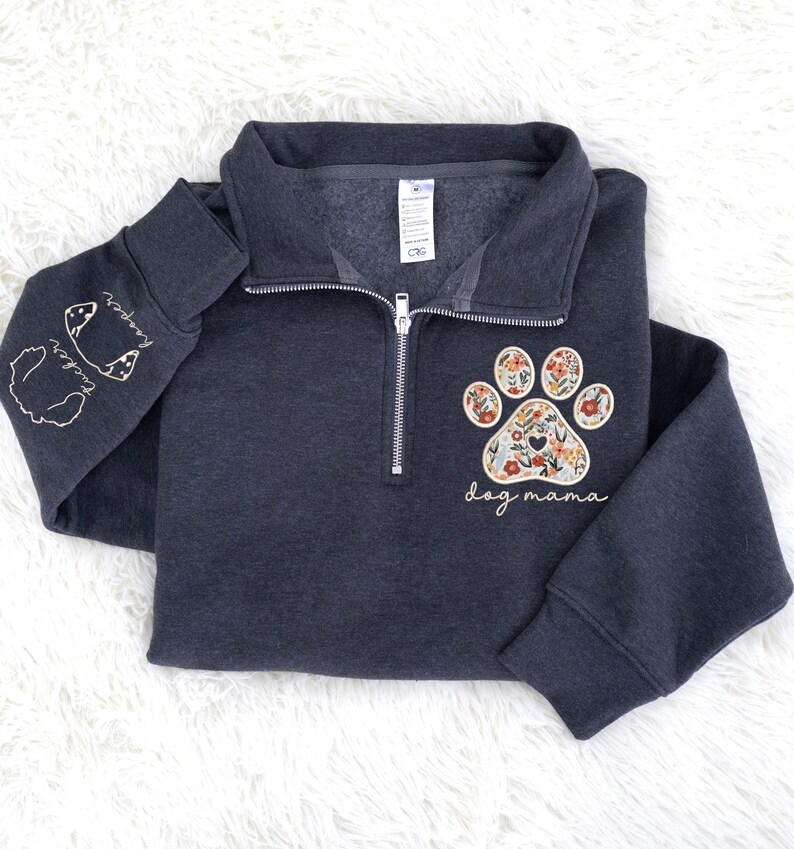 May include: Dark gray quarter-zip sweatshirt with a floral paw print design and the words "dog mama." The left sleeve features embroidered dog silhouettes and names. The sweatshirt has a silver zipper and a soft, comfortable appearance.