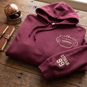 May include: A maroon hooded sweatshirt featuring an embroidered design on the chest with a football graphic and the text "football mom". The sleeve is embroidered with the name "ETHAN" and the number "99" in a light tan thread color.