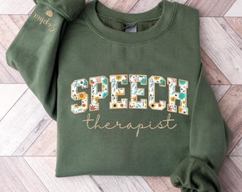 May include: A dark green sweatshirt with the words "SPEECH therapist" in gold lettering. The word "SPEECH" is decorated with colourful floral patterns.