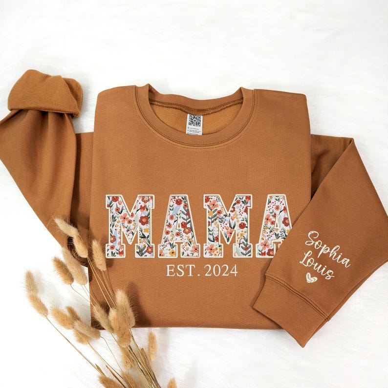 May include: A brown sweatshirt with the word "MAMA" in large, floral-filled letters. Below the word "MAMA" is the text "EST. 2024". The sleeve has the embroidered name "Sophia Louis" with a small heart. The sweatshirt is a cozy, casual style.