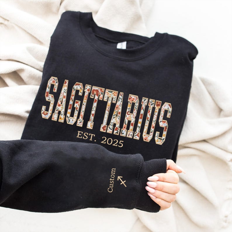 May include: Black sweatshirt with a floral Sagittarius design and the text "EST. 2025". The sleeve has a small Sagittarius symbol and the word "Custom" in gold.
