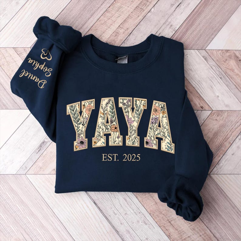 May include: Navy blue sweatshirt with the word "YAYA" in a floral design. The text "EST. 2025" is below the word. The left sleeve has the names "Daniel" and "Sophia" embroidered in gold.