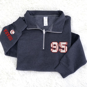 May include: A dark gray baseball-themed pullover sweatshirt with a quarter-zip neckline. The number "95" is embroidered on the front in red and white, with baseball stitching. The left sleeve has the name "SHARA" and a baseball embroidered in red.
