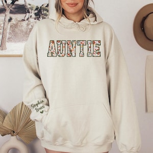 May include: Cream-colored hoodie with the word "AUNTIE" in a floral design across the chest. The sleeve features the name "Sophia Louis" in script. The hoodie has a drawstring hood and a front pocket.