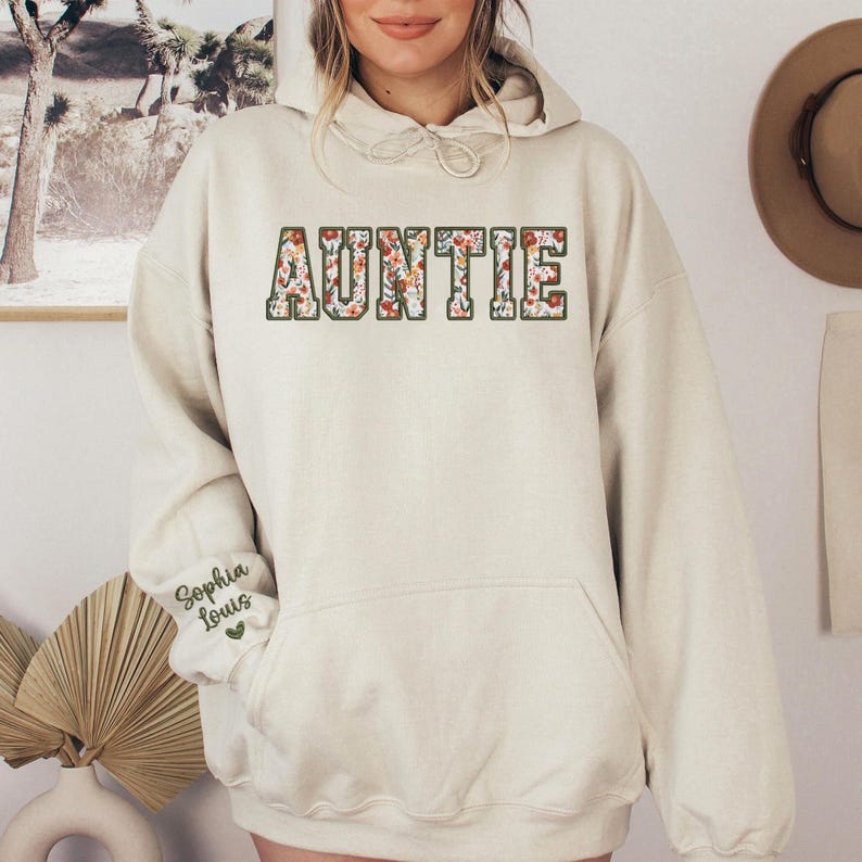 May include: Cream-colored hoodie with the word "AUNTIE" in a floral design across the chest. The sleeve features the name "Sophia Louis" in script. The hoodie has a drawstring hood and a front pocket.