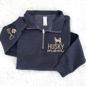 Op de afbeelding: Donkergrijze sweatshirt met een kwart rits en gouden borduursels. Het ontwerp bevat een husky-hondsilhouet en de woorden "HUSKY mom". De linkermouw heeft de naam "Daisy" en een voetafdruk.