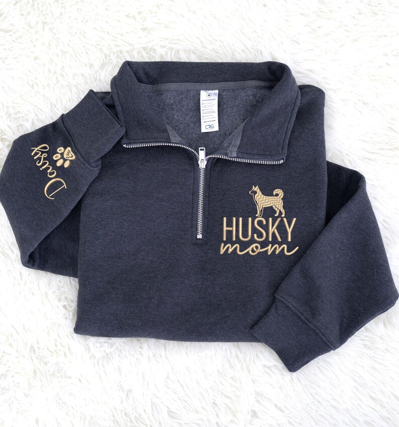 Op de afbeelding: Donkergrijze sweatshirt met een kwart rits en gouden borduursels. Het ontwerp bevat een husky-hondsilhouet en de woorden "HUSKY mom". De linkermouw heeft de naam "Daisy" en een voetafdruk.
