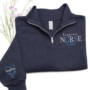 May include: Navy blue quarter-zip sweatshirt with the words "hospice NURSE" embroidered in white and light blue. The sleeve features a heart-shaped stethoscope design with the name "Sophia" embroidered in white and light blue. The sweatshirt has a silver zipper.