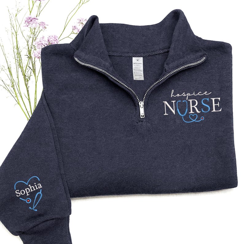 May include: Navy blue quarter-zip sweatshirt with the words "hospice NURSE" embroidered in white and light blue. The sleeve features a heart-shaped stethoscope design with the name "Sophia" embroidered in white and light blue. The sweatshirt has a silver zipper.