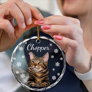 May include: A round, clear glass ornament with a gold string for hanging. The ornament features a brown tabby cat with green eyes and the name "Chopper" printed in black. The background is a dark blue with white snowflakes.