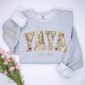 May include: A light gray sweatshirt with the word "YAYA" in a floral design. The sweatshirt has the text "EST. 2025" below the word "YAYA". The left sleeve has the names "Daniel Sophia" embroidered in gold.