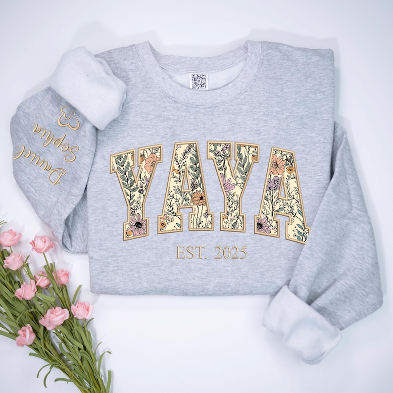 May include: A light gray sweatshirt with the word "YAYA" in a floral design. The sweatshirt has the text "EST. 2025" below the word "YAYA". The left sleeve has the names "Daniel Sophia" embroidered in gold.