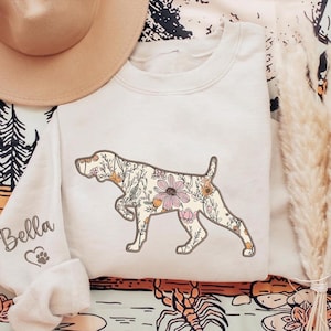 May include: A white sweatshirt with a floral print of a dog silhouette. The dog is outlined in brown and the flowers are in shades of pink, orange, and yellow. The text "Bella" is embroidered on the sleeve of the sweatshirt.
