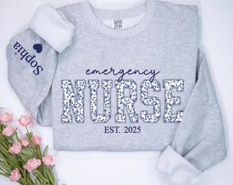 May include: A gray sweatshirt with a floral pattern spelling "emergency nurse" and the text "EST. 2025". The sweatshirt has a white cuff with the name "Sophia" embroidered on it.