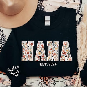 May include: Black sweatshirt with the word "NANA" in large, floral-patterned letters. Below the word "NANA" is the text "EST. 2024". The sleeve has the text "Sophia Louis" and a heart. A tan hat is in the background.