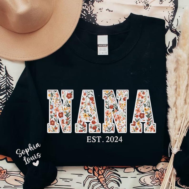 May include: Black sweatshirt with the word "NANA" in large, floral-patterned letters. Below the word "NANA" is the text "EST. 2024". The sleeve has the text "Sophia Louis" and a heart. A tan hat is in the background.