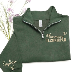 May include: A dark green quarter-zip sweatshirt with the words "Pharmacy Technician" embroidered in a gold color. The name "Sophia" is embroidered on the sleeve. The sweatshirt has a silver zipper and a small heart design.