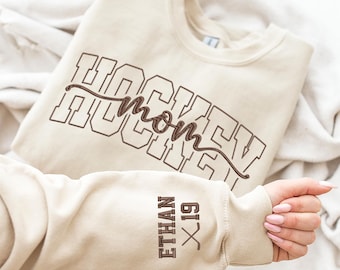 May include: A beige sweatshirt with the embroidered text "HOCKEY mom" in brown. The sleeve features the name "ETHAN" and the number "19" in brown embroidery. The sweatshirt is folded on a white surface.