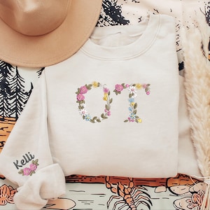 May include: Cream-colored sweatshirt with floral embroidered letters "OT". The letters are adorned with pink, yellow, and white flowers. The left sleeve has the name "Kelli" embroidered with a floral design. A tan hat is visible in the upper left corner.