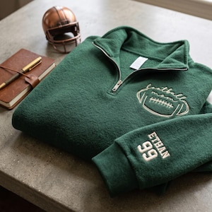 May include: A dark green quarter-zip pullover featuring white embroidery on the chest with a football graphic and the text "football mom". The sleeve is embroidered with the name "ETHAN" and the number "99" in white block lettering.