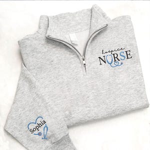 May include: A light gray quarter-zip sweatshirt with the words "hospice NURSE" embroidered in black and blue. The sleeve features the name "Sophia" and a stethoscope design. The sweatshirt has a silver zipper.