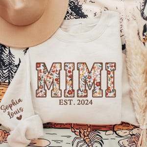 May include: A white sweatshirt with the word "MIMI" in brown letters with a floral pattern inside. The text "EST. 2024" is below the word "MIMI".