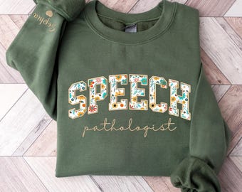May include: A green sweatshirt with the words "SPEECH pathologist" in gold lettering. The word "SPEECH" is decorated with a floral pattern.