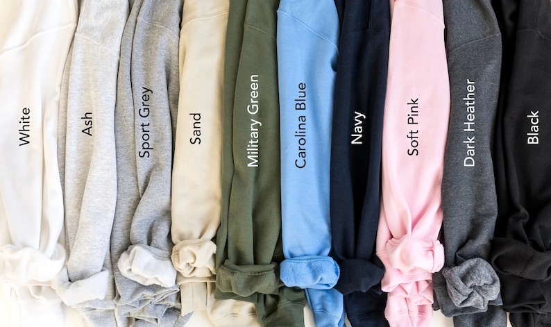 May include: A collection of ten sweatshirts in various colors, including white, ash, sport grey, sand, military green, carolina blue, navy, soft pink, dark heather, and black. Each sweatshirt is displayed with its color name.