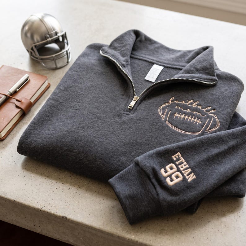 May include: A charcoal gray quarter-zip pullover featuring rose gold embroidery on the chest that reads football mom with a football graphic. The sleeve is embroidered with the name ETHAN and the number 99 in matching rose gold thread.